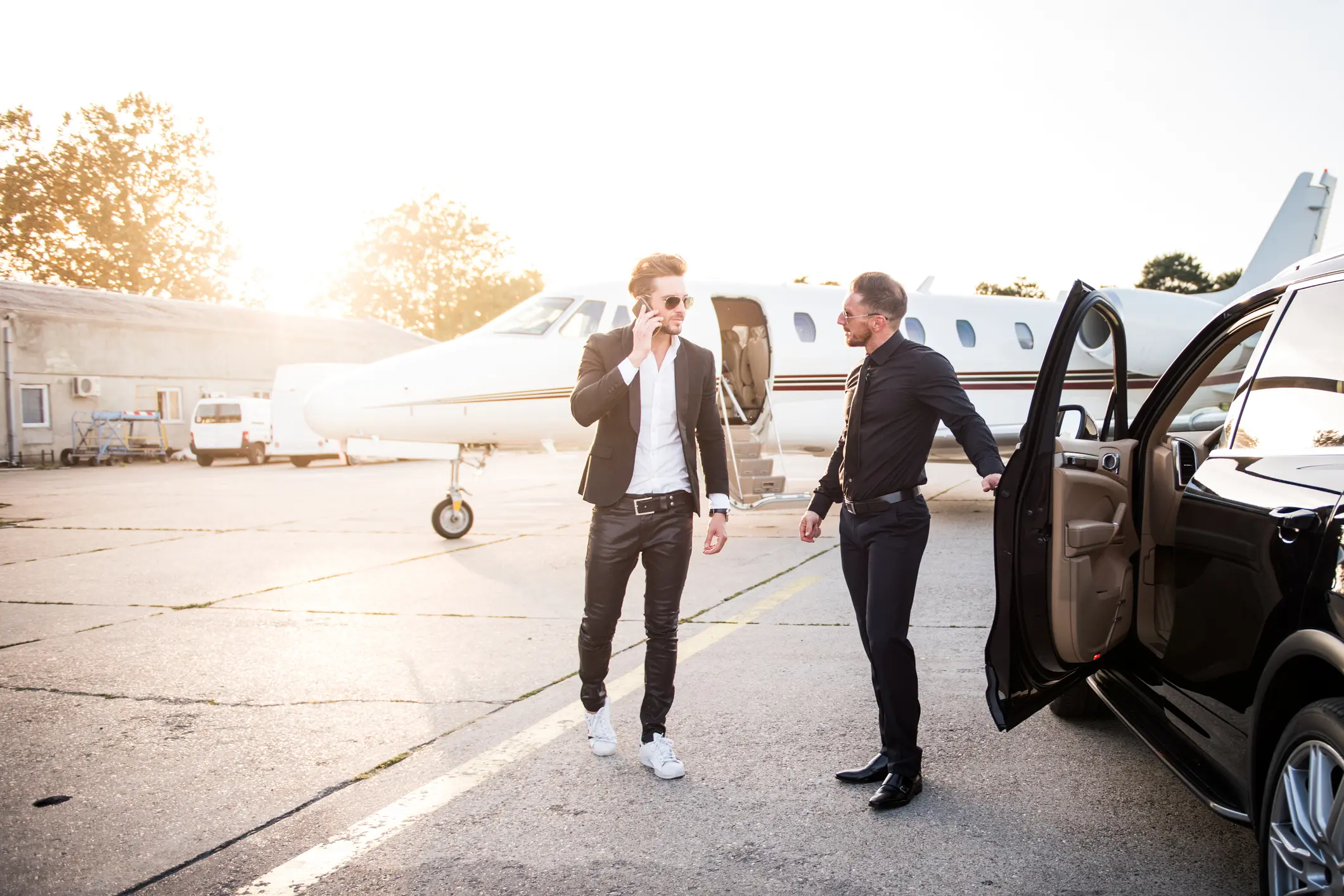Man wearing black clothes and sunglasses standing next to a car and welcoming another man who arrived by private jet airplane.